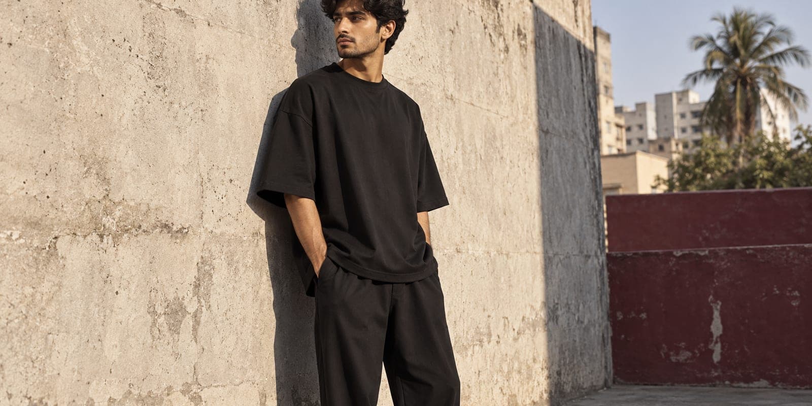 A young man in a plain black oversized drop-shoulder t-shirt standing against a sunlit concrete wall in Karachi